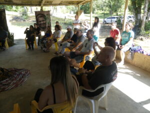 Group of people sitting in an outdoor patio space. They are sitting in yellow chairs in a semi circle and some are standing behind the seated people.