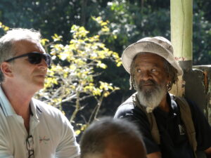 Two men sitting together with greenery in the background. One man is wearing a hat and looking at the other man wearing sunglasses.