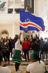 Audience of people standing and watching woman wave Republic of Cape Verde flag. She is wearing a green skirt and green headband.