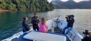 Three people having a conversation on a boat in the water being filmed by a camera person.