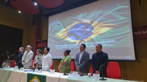 Group of speakers behind a table with a presentation of Brazilian flag behind them.