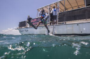 A man is stepping off a boat named the "Cheeca View." The man wears a oxygen tank, diving fins and goggles. Behind him a man looks over the boat watching him jump. There is a sign next to the second man reading, "Horizon Divers." the ocean water takes up the bottom 1/4 of the frame.