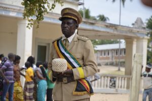 a man in a uniform holding a basket made of shells. in the background there is a large building and group of people looking behind them.
