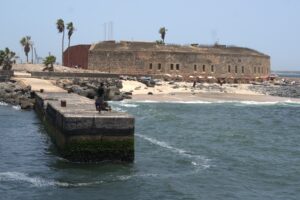 a landscape ofa beach. there is a person standing on a stone dock towards the foreground. in the background is a brick building and a stone-covered beach.