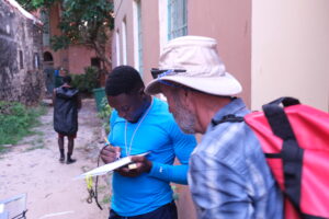 Man taking notes while another man speaks to him, carrying a large backpack, in an alleyway.