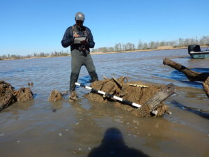 A man stands in the middle of the frame, looking down at a clipboard. He stands in water up to knees. In the fore ground are several pieces of wood sticking up out of the water. On the largest piece of wood, sits two measuring devices. On the right side of the frame a boat's motor is visible. The background is divided between blue sky and brown water but a line of trees.