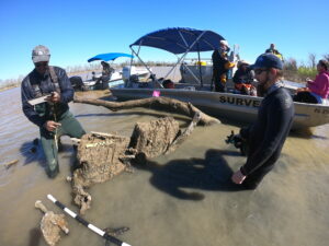 Two men stand in the foreground, both are in wetsuits with water up to their knees. They are examining pieces of wood protruding up out of the water. the largest piece of wood has a pink flag sticking out of it and measuring equipment nearby. One man examines a clipboard. Behind them are two small boats with five other people visible on them.