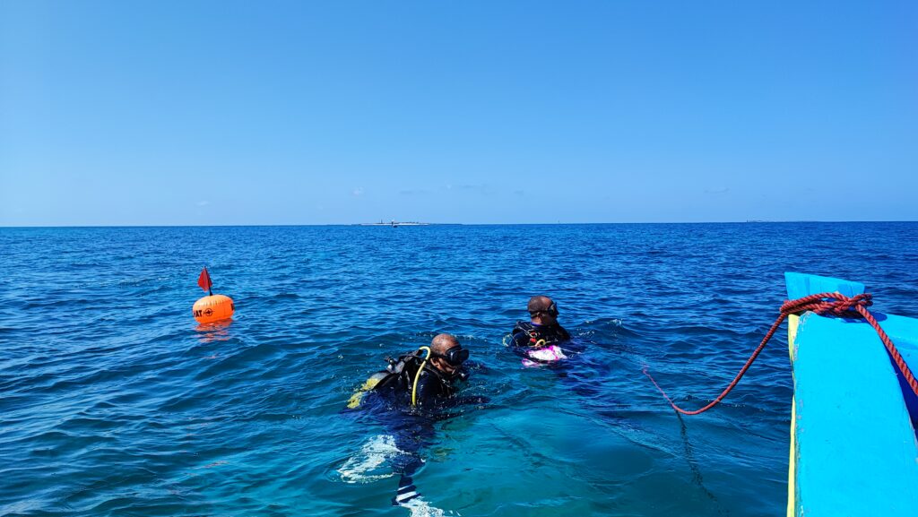 two people in scuba gear floating at the surface of the water. their shoulders and heads are still above the water but their legs are submerged. the front of a boat is cut off in the forground, with a rope that is descended towards the divers.