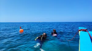 two people in scuba gear floating at the surface of the water. their shoulders and heads are still above the water but their legs are submerged. the front of a boat is cut off in the forground, with a rope that is descended towards the divers.