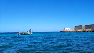 landscape of the ocean. on the left edge, there are two small boats with a large group of people standing on them. on the right side, there is a large stone building.