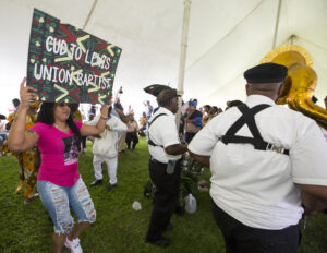 Woman holding up a sign that reads "Cudjo Lewis Union Baptist." In front of her men holding drums face away towards the rest of a band. Behind the woman other people march in a circle. Everyone is under a white tent.