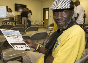 A man wearing a yellow shirt and black and white hat looks towards the camera. He is sitting in a metal chair arranged for a presentation. People in the background stand around the presentation area. the man is holding a piece of paper to the camera reading, "Clotilda Discovery Celebration!! We Found it!"