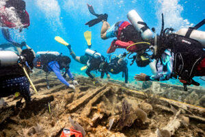 Large group of divers investigating wreckage on ocean floor.