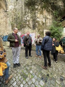 Group of people standing together in conversation. They are on a coblestone street with a big building with vines behind them.