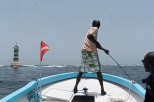 a person standing at the front of a boat, pulling on rope. there is a lighthouse in the background on top of the ocean water.