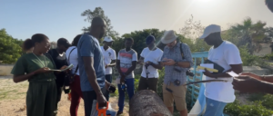 A group of people standing and looking at a canon together outside. On the far right, a white man with a grey beard is wearing a tan hat, sunglasses, a blue plaid shirt with the sleeves rolled up, and khaki pants. Other men and women of African descent stand around looking at the canon, some holding notepads, cameras, or phones in their hands. In the background, trees and vegetation is visible, as well as some buildings barely visible in the distance. The people appear to be talking and taking notes.