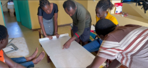 A group of people sitting around a table and standing, looking at a map. The person in the center is pointing to a point in the map. He wears a dark green shirt and navy shorts, and has short hair. On either side of him are two women. To his left is a woman with long dreads tied back wearing a yellow shirt and bluejeans, along with square earrings. The woman on his right wears a dark navy top and lighter blue skirt with buttons down the front. In the bottom right foreground, a person wears a brown, tan, and white striped shirt. They have short hair and their face is not visible. The room they are in is yellow with tiled floors.