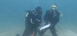 Two divers underwater in scuba gear. One is holding a clipboard. They both have short hair and dark skin. The diver on the left holds the clipboard and is writing. He wears a black dive mask and has on a dark colored scuba suit with white writing on the sleeve. The diver on the right holds up his left hand, making a signal. He wears a dark colored scuba suit with long sleeves, and a yellow scuba mask.