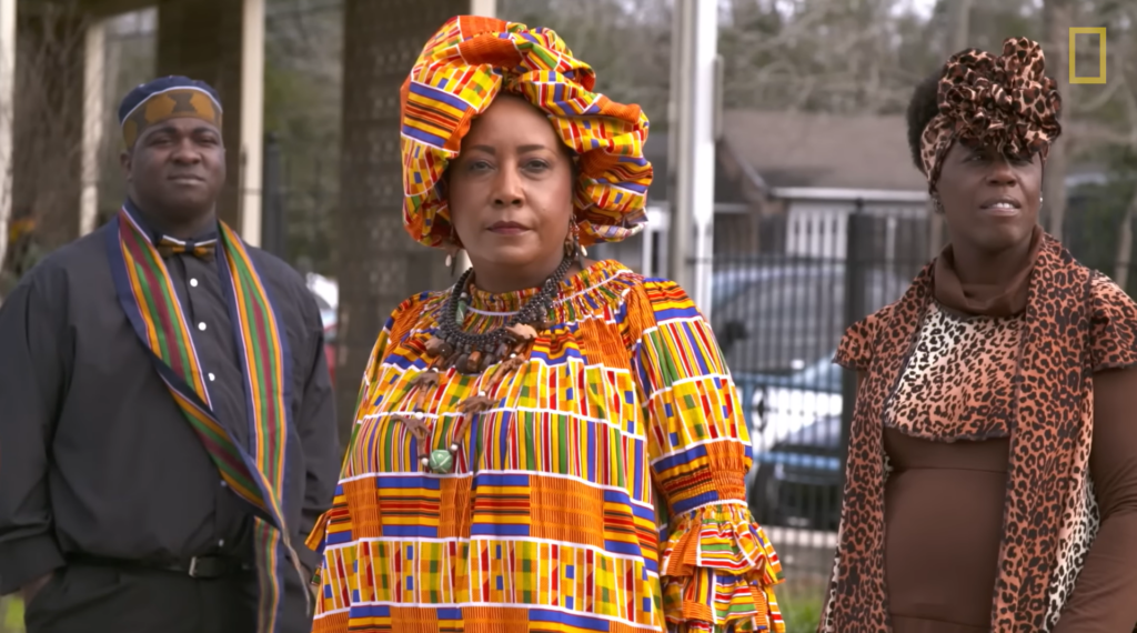 Three people stand in frame, the two on the sides of the center look into the distance while the person in the center looks into the camera. All three people have dark skin. The central person wears a brightly colored orange, blue, green, yellow, and red patterned cloth dress and matching hat. They wear a large necklace around their neck that is black and has beads. The person on the right wears a brown dress with a leopard print shawl, and a matching leopard bring head band with a bow. The person on the left wears a black collared shirt with a blue, orange, red, and white bowtie and matching scarf. They wear a hat that is blue, brown, and white.