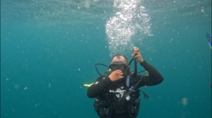 a diver underwater holding their nose with their hand. there are bubbles coming up from him.