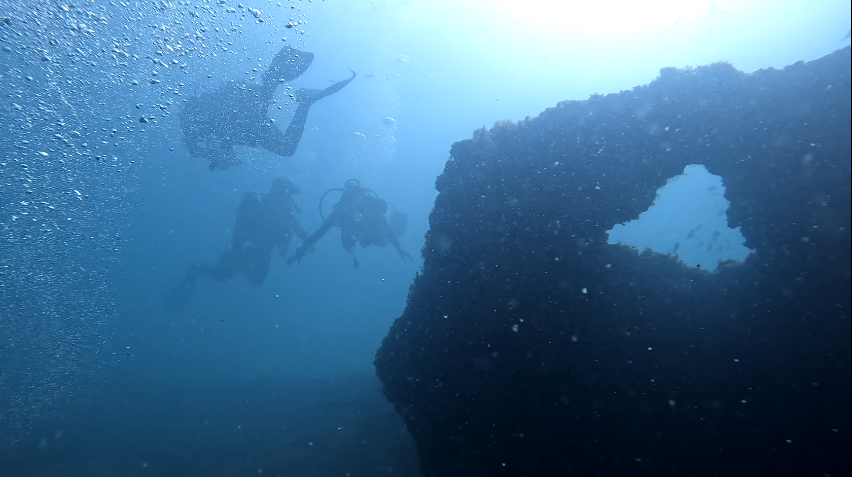 Three scuba divers swim in deep water. They are wearing scuba diving gear, but their faces are not visible. In the foreground, a large circular structure is visible. The water is deep blue and a few bubbles are visible in the foreground.