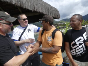 Four men are visible in the photo. Two of them are shaking hands while talking to the third man. The fourth man smiles at the other three. Behind them a straw roof is visible. Behind the some buildings an water are visible. The background, green hills rise to meet the grey sky.