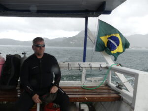 A man sits on a bench aboard a boat. He is dressed in a wetsuit and sunglasses and is looking at the water. Next to him a Brazilian flag waves in the winds, under the boats awning. In the background, rough waters are visible. Behind them is a mount shrouded in grey skies and several dark clouds. The slight blurriness of the photo suggests that rain drops have hit the camera lens.