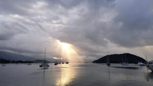 The image is of a cove with several sail boats anchored in the water. In the background several hills rise up out of the water. The sky is made of dense grey clouds, expect for in the center of the frame where rays of sunlight shine through a small opening.