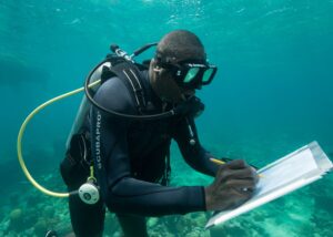 Black man in scuba gear underwater, taking notes on a laminated sheet.