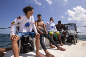 Four men sit on a bench on a boat. They are wearing a variety of swimming gear including swim trunks and wetsuits. The mean are talking and looking at theirs phones. Behind them the blue ocean is visible along with a blue sky marked by several white clouds.