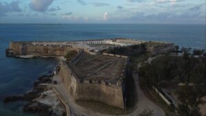 The image shows an old stone fort. It is areial view showing that fort has both sea and land walls. in the center for the fort, white buildings are visible but look abandoned. the fore ground is the road leading to the fort and surround trees. The background is split between blue ocean and cloudy skies.