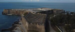 The image shows an old stone fort. It is areial view showing that fort has both sea and land walls. in the center for the fort, white buildings are visible but look abandoned. the fore ground is the road leading to the fort and surround trees. The background is split between blue ocean and cloudy skies.