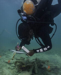 A diver examines the ocean floor. He is wearing full scuba gear, including goggles, a wetsuit and oxygen tank. He holds a clipboard and is writing on it with a pencil. Below him the ocean floor is visible, on it a pile of wood with two orange flags sticking out.