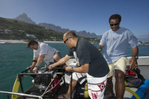 Three men stand on a boat. Two are pulling up a rope attached to larger tube. The third man walks behind them and is wearing a blue shirt with "Dive" written on it. None are looking at the camera. Scuba equipment can be seen behind them. In the Background blue water meets a green shoreline before rising into small mountains/