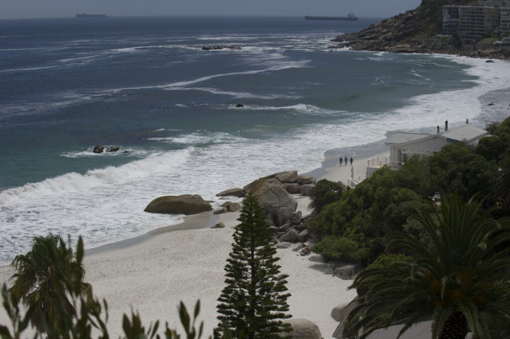 A shoreline takes up the upper left half of the frame and the lower right half is covered in beech, green trees and one house. In the distance rocks move out form the shoreline creating a small cove. On the horizon a large freight ship is visible. Three people walk on the shore.