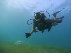 A diver underwater is looking at the camera, holding up a thumbs up. The diver is wearing gloves, a oxygen tank and diving fins. Behind him, the grassy ocean floor is visible with a yellow flag sticking out of the ground.