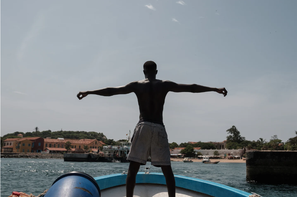 Black man stands at the front of a boat, which is cut off in in the framew with arms outstretched besides him. There is a beach and buildings in the background behind the waves.