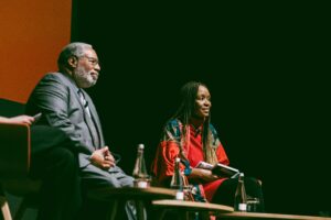 Two people sitting on stage listening to another speaker. A man sits on the left. He has a short grey beard and short curly gray hair. He is wearing a gray suit. The woman on the right has long braids, and is wearing a red and blue top. She holds a notepad in her hand. In front of them, there is a table with several bottles of water.