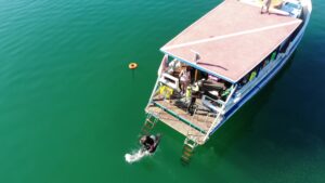 The frame is taken up by an overhead shot of a boat. with a red and white roof. Off the back of the boat a person in full dive gear jumps into calm green waters. Another people in dive gear stands on the boats edge.