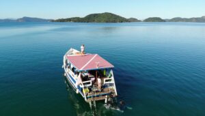 In the foreground a boat sits in clam waters. A man stands on the boats roof. Off the back of the boat, two people are in the water, dressed in but scuba gear. the majority of the frame is taken up with calm water. At the top of the frame, small hills are visible before a clear blue sky.