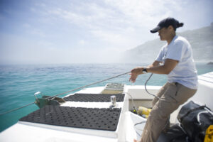 a person standing at the edge of a boat pulling rope. the rope is coming from the water on the edge. behind them is mountain and more ocean.