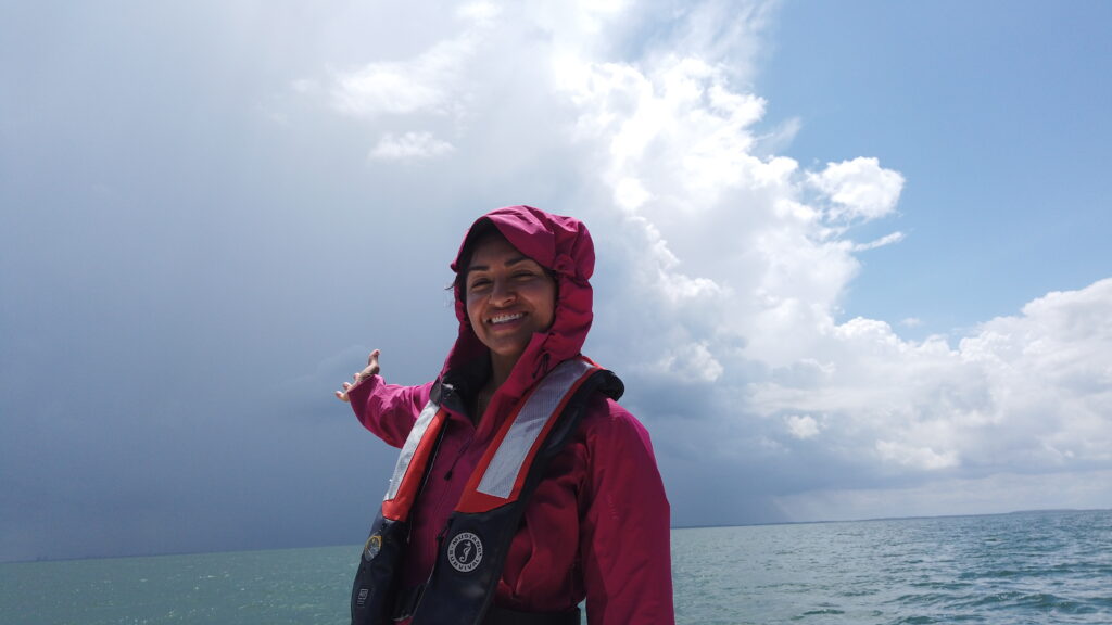 a girl stands and points behind her, smiling at camera. behind her is the ocean with big clouds covering the sky.