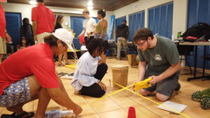 a group of three practices using measuring tapes on tile floor inside, kneeling on the ground together measuring. a group is behind them talking and looking at another measuring tape.