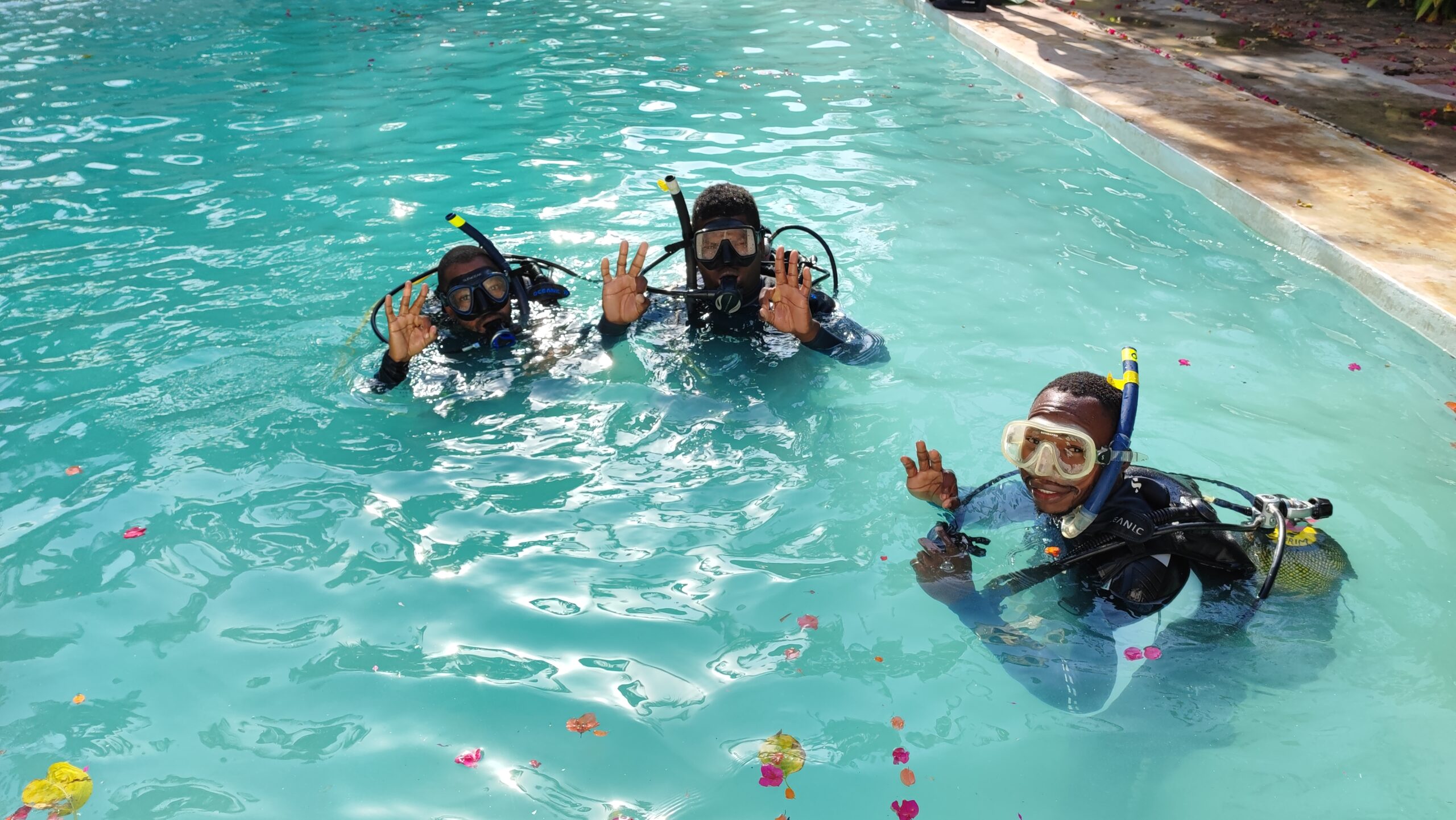 Three divers in a pool smiling and posing. They are visible from the neck up, and each wears a scuba mask and snorkel.