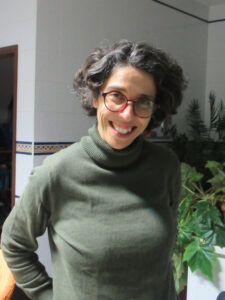 Woman smiling in a room, her shoulders and chest are visible with green plants behind her. She wears a dark green turtleneck sweater, red glasses, and has a short wavy bob hairstyle.