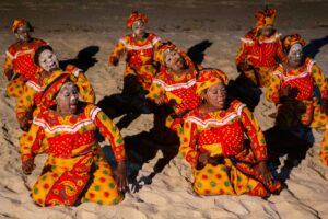 Women sitting in sand singing and dancing in ceremonial dress.