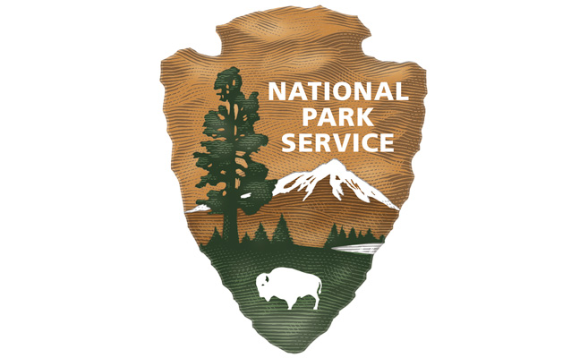 National Parks Services Logo. The logo is in the shape of an arrow head and has sketchy lines to give it dimensionality. There is a large tree in the foreground and smaller conifer trees in the background, as well as a snow capped mountain. Above the mountain, it says "NATIONAL PARK SERVICE" in white block font. At the bottom of the arrowhead, there is a white buffalo figure standing on a green background.