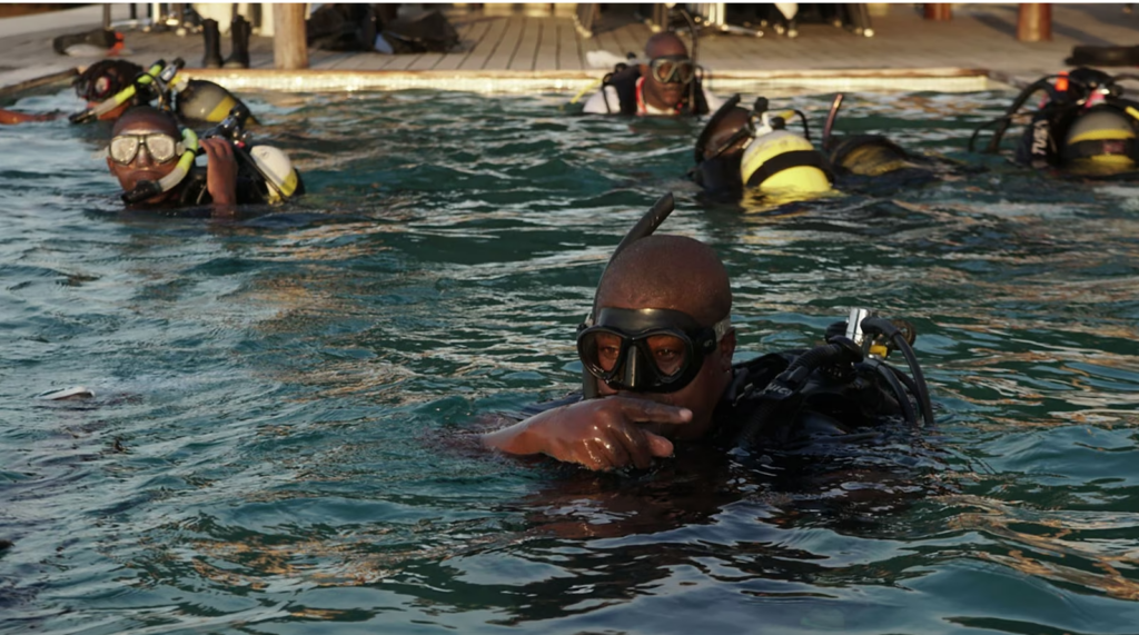 Group of divers in the water with their heads out of the water on the surface. They are swimming in a swimming pool. They all have dark skin. One diver is closest to the photographer, and has a bald head and is wearing a black dive mask and snorkel. He is pointing to his left. In the background, a wooden porch is visible.