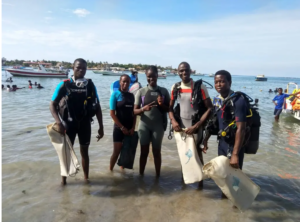 Group of five divers of African descent standing in water up to their ankles. There are boats and other groups of people behind them in the water. They are wearing gear and smiling together. A bar of land is visible in the background.