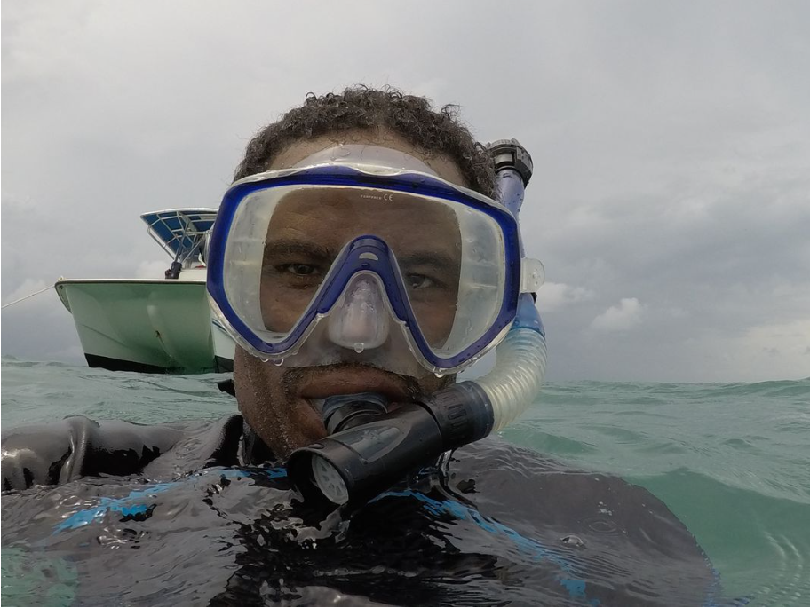 Man taking photo in the water, his shoulders are underwater and he is wearing goggles and scuba gear. There is a boat behind him in the water. He has dark skin and is wearing a dark colored scuba top. He has short hair and slightly scruffy facial hair.